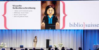Das Bild zeigt Saphir Ben Dakon auf der grossen Bühne beim Kongress von Bibliosuisse. Hinter ihr ist eine Folie eingeblendet, in der ihre visuelle Selbstbeschreibung nachzulesen ist. 