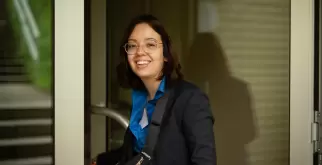 A picture of Saphir Ben Dakon looking sideways into the camera and laughing. She is opening a door. She is wearing her golden glasses, a dark blue blouse and a matching dark blue blazer.