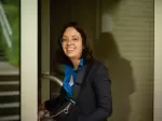 A picture of Saphir Ben Dakon looking sideways into the camera and laughing. She is opening a door. She is wearing her golden glasses, a dark blue blouse and a matching dark blue blazer.