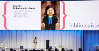 Das Bild zeigt Saphir Ben Dakon auf der grossen Bühne beim Kongress von Bibliosuisse. Hinter ihr ist eine Folie eingeblendet, in der ihre visuelle Selbstbeschreibung nachzulesen ist. 
