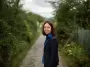 Saphir Ben Dakon stands on a gravel path on the Hönggerberg. She looks at the camera and laughs. Behind her is green and the path can be identified. She is wearing her gold glasses, a blue blouse and a matching blue blazer.