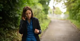 Saphir Ben Dakon am Telefon. Sie geht dabei einen Waldweg entlang und das Gesicht ist der Kamera abgewandt. Sie trägt eine blaue Bluse und einen passenden blauen Hosenanzug dazu.