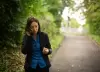 Saphir Ben Dakon am Telefon. Sie geht dabei einen Waldweg entlang und das Gesicht ist der Kamera abgewandt. Sie trägt eine blaue Bluse und einen passenden blauen Hosenanzug dazu.